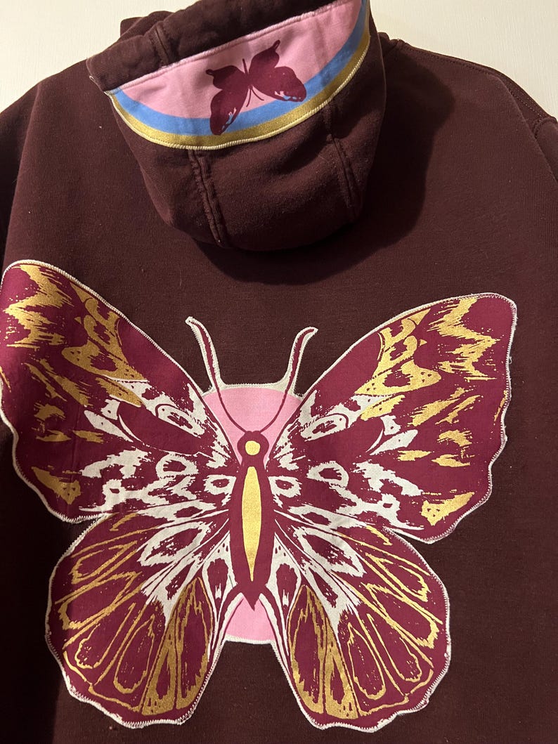 Puede incluir: Una sudadera con capucha granate con un gran dise&ntilde;o de mariposa en la espalda. La mariposa tiene detalles dorados y blancos. La capucha tiene un gr&aacute;fico de mariposa rosa y rayas azules y amarillas.