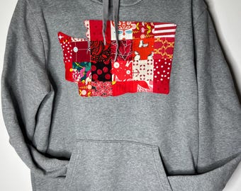 May include: A heather gray hooded sweatshirt featuring a large, central applique in the shape of Washington state. The applique is constructed from a patchwork of various red, white, and black fabrics with diverse patterns, including polka dots and florals.