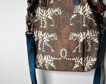 May include: A brown tote bag with a repeating pattern of white tigers. The bag has brown leather handles and a detachable blue floral patterned shoulder strap. The bag is made of a sturdy fabric.