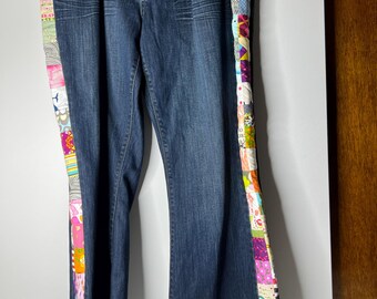 May include: Dark wash denim jeans with colorful patchwork accents down the sides. The patchwork features various patterns and colors, including pink, red, green, and purple. The jeans are hanging on a hanger.