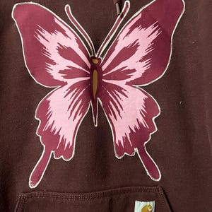 Puede incluir: Sudadera con capucha granate con un gran dise&ntilde;o de mariposa. La mariposa tiene alas rosas y granates con contornos blancos. La sudadera tiene un bolsillo de canguro y una etiqueta Carhartt.