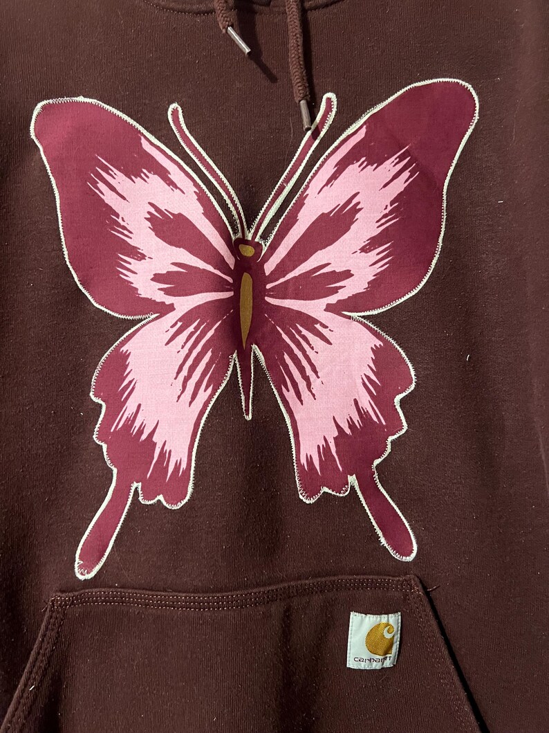 Puede incluir: Sudadera con capucha granate con un gran dise&ntilde;o de mariposa. La mariposa tiene alas rosas y granates con contornos blancos. La sudadera tiene un bolsillo de canguro y una etiqueta Carhartt.
