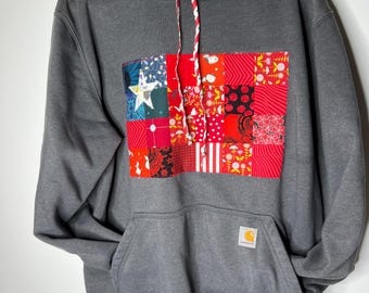 May include: A charcoal gray hooded sweatshirt featuring a large rectangular patchwork panel on the chest composed of various red, black, and white patterned fabrics, including a white star, red and white drawstrings, and a Carhartt brand patch.