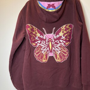 Puede incluir: Sudadera con capucha burdeos con un gran dise&ntilde;o de mariposa en la espalda. La mariposa es rosa, dorada y blanca. La capucha tiene un acento rosa y azul con un dise&ntilde;o de mariposa m&aacute;s peque&ntilde;o.