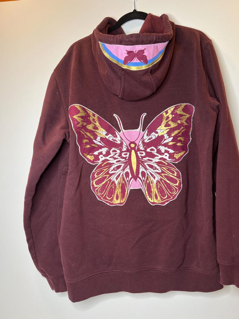 Puede incluir: Sudadera con capucha burdeos con un gran dise&ntilde;o de mariposa en la espalda. La mariposa es rosa, dorada y blanca. La capucha tiene un acento rosa y azul con un dise&ntilde;o de mariposa m&aacute;s peque&ntilde;o.