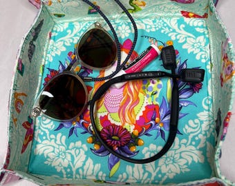 May include: A square fabric tray with a colorful floral and paisley pattern. The tray contains sunglasses, a lip balm, and a black cord. The interior fabric is turquoise with white floral designs. The tray's edges are multi-colored.