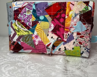 May include: A rectangular zippered pouch featuring a patchwork design made of various fabric scraps in diverse colors and patterns, including floral, geometric, and solid prints, all held together with visible white grid-style machine quilting.