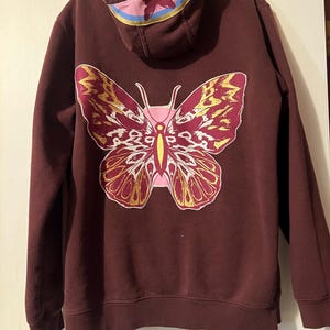 Puede incluir: Una sudadera con capucha granate con un gran dise&ntilde;o de mariposa en la espalda. La mariposa tiene detalles rosas, amarillos y blancos. La capucha tiene una franja rosa y azul con un gr&aacute;fico de mariposa. La sudadera es de material suave.