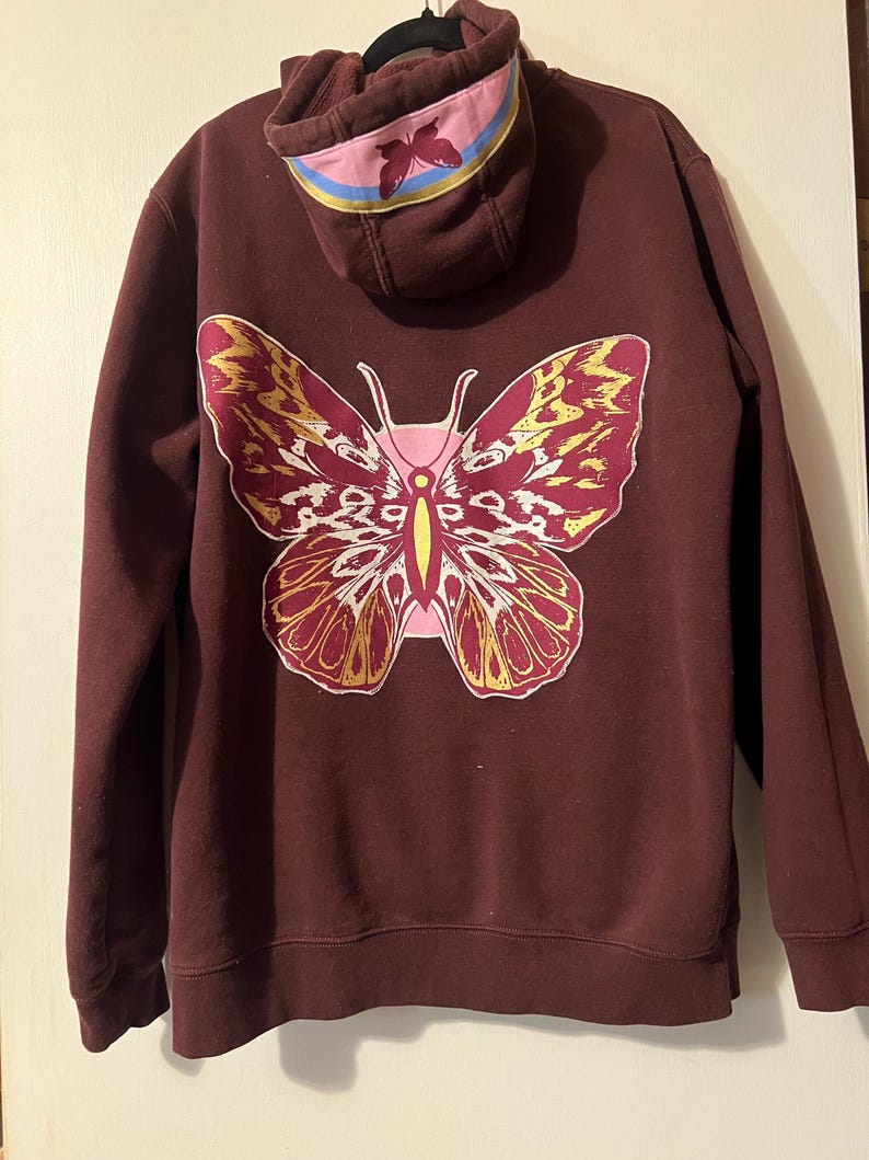 Puede incluir: Una sudadera con capucha granate con un gran dise&ntilde;o de mariposa en la espalda. La mariposa tiene detalles rosas, amarillos y blancos. La capucha tiene una franja rosa y azul con un gr&aacute;fico de mariposa. La sudadera es de material suave.