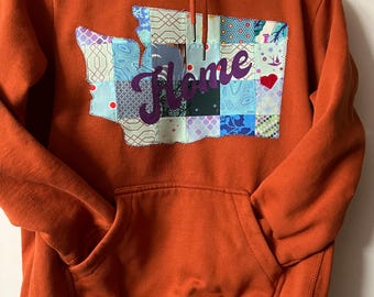 May include: A burnt orange hooded sweatshirt featuring a large, central patch shaped like the state of Washington. The patch is constructed from various blue, white, and patterned fabric squares and displays the word Home in purple script.