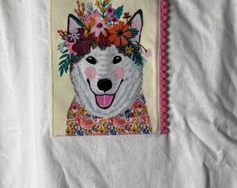May include: White t-shirt with a patch featuring a smiling husky wearing a floral crown and a floral patterned collar. The patch is bordered with pink pom-poms.