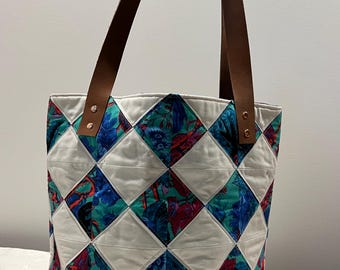 May include: A quilted tote bag with a diamond pattern. The bag features a white background with teal, blue, and red floral fabric in the diamond shapes. It has brown leather handles attached with metal rivets.