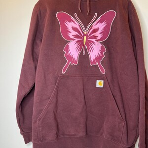 Puede incluir: Sudadera con capucha burdeos con un gran dise&ntilde;o de mariposa rosa y magenta en la parte delantera. La sudadera tiene un bolsillo de canguro y un peque&ntilde;o parche con el logotipo de Carhartt. La capucha tiene cordones con ojales met&aacute;licos dorados.