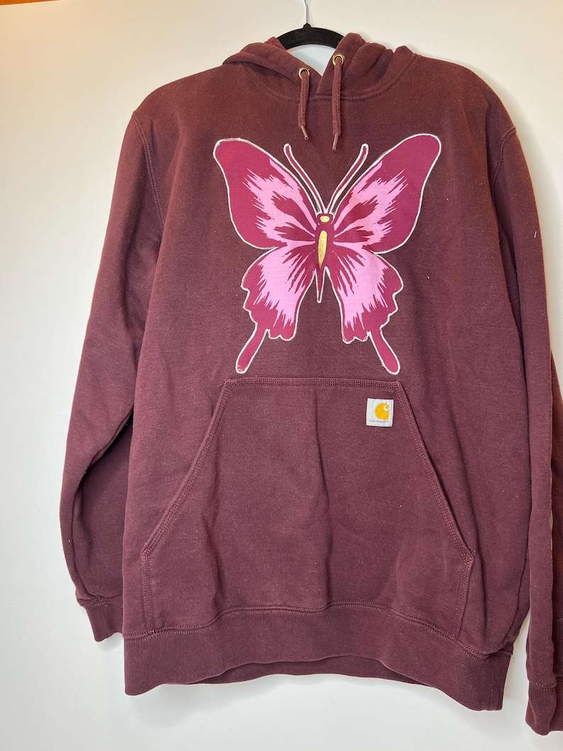 Puede incluir: Sudadera con capucha burdeos con un gran dise&ntilde;o de mariposa rosa y magenta en la parte delantera. La sudadera tiene un bolsillo de canguro y un peque&ntilde;o parche con el logotipo de Carhartt. La capucha tiene cordones con ojales met&aacute;licos dorados.