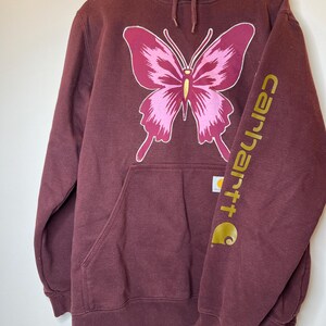 Puede incluir: Sudadera con capucha burdeos con un gran dise&ntilde;o de mariposa rosa. El logotipo de Carhartt est&aacute; impreso en dorado en la manga. Cuenta con un bolsillo delantero y una capucha con cord&oacute;n. Una sudadera informal y c&oacute;moda.