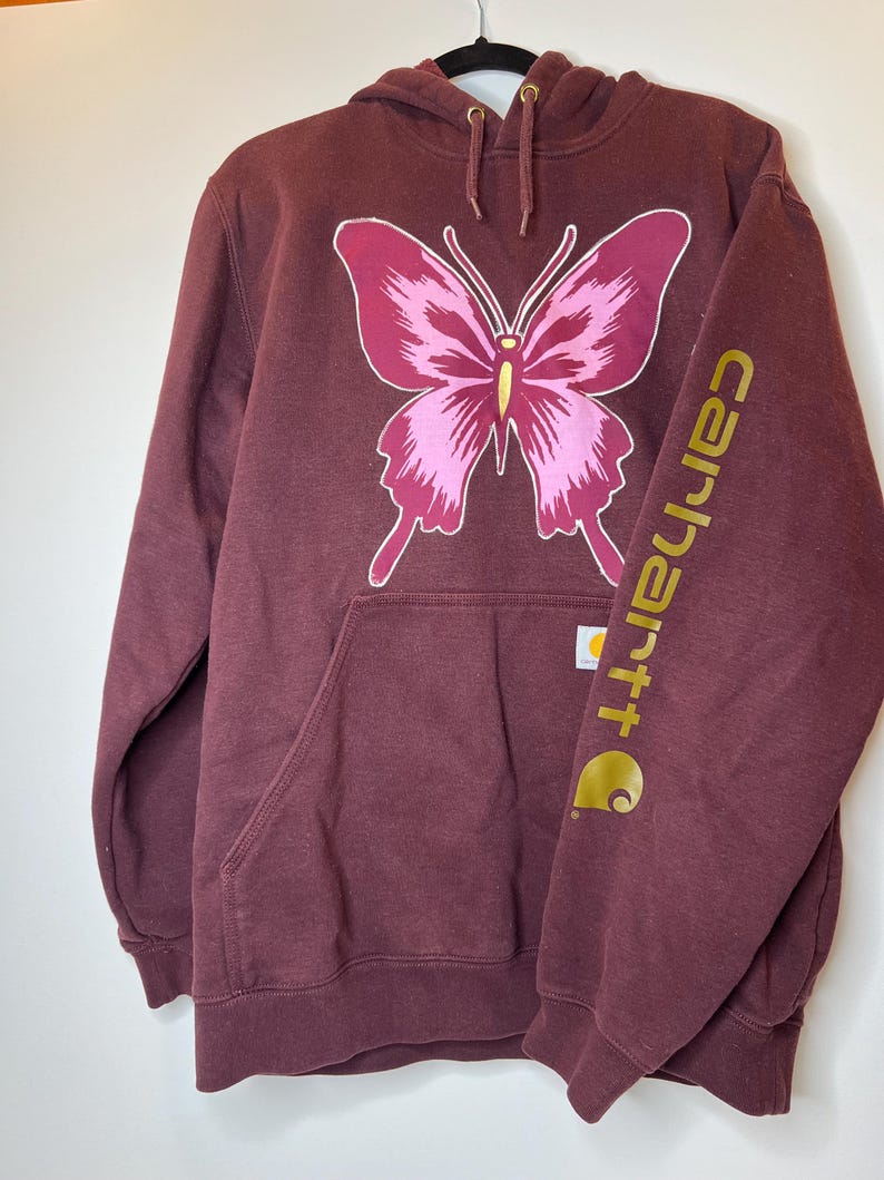 Puede incluir: Sudadera con capucha burdeos con un gran dise&ntilde;o de mariposa rosa. El logotipo de Carhartt est&aacute; impreso en dorado en la manga. Cuenta con un bolsillo delantero y una capucha con cord&oacute;n. Una sudadera informal y c&oacute;moda.