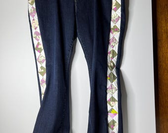 May include: Dark wash denim jeans with a flared leg. The jeans feature a decorative patchwork stripe down the side of each leg, with a pattern of pink, green, and yellow floral and geometric shapes. The jeans are hanging on a hanger.