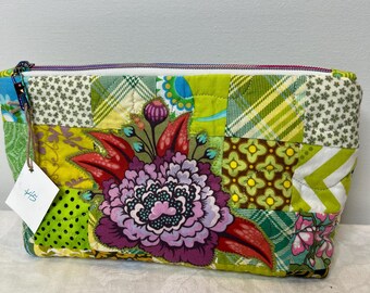 May include: A rectangular quilted zipper pouch featuring a patchwork design of green, yellow, and white fabrics with various patterns. A large, vibrant floral applique in shades of purple, red, and green is centered on the front. A white tag is attached.