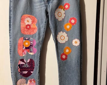 May include: Light blue denim jeans with decorative floral and letter patches. The left leg features the letters "BOHO" in colorful patchwork. The right leg has embroidered flower appliques in shades of white, orange, and pink.