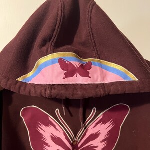 Puede incluir: Una sudadera con capucha granate con un gran gr&aacute;fico de mariposa en la parte delantera. La capucha presenta un dise&ntilde;o de mariposa m&aacute;s peque&ntilde;o y un acento de arco&iacute;ris colorido. Los dise&ntilde;os de mariposas son en tonos rosa y granate.