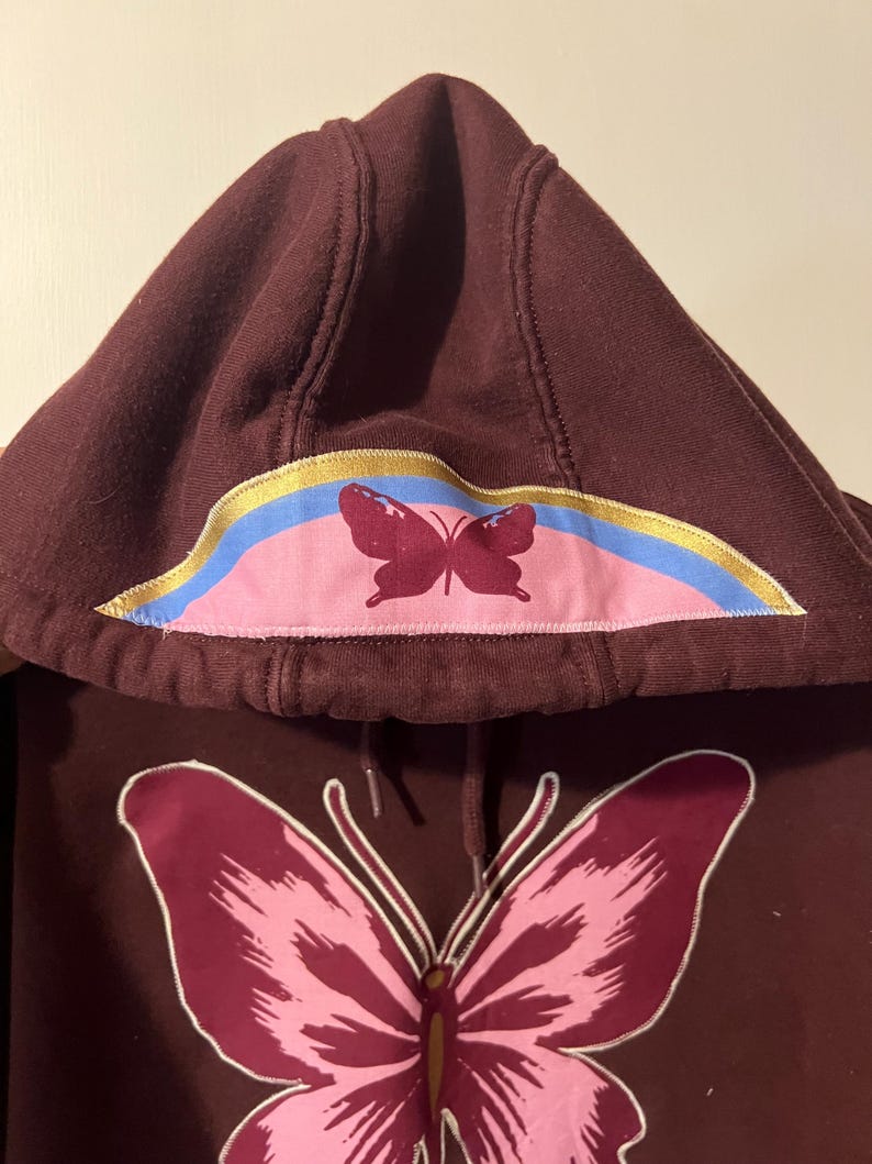 Puede incluir: Una sudadera con capucha granate con un gran gr&aacute;fico de mariposa en la parte delantera. La capucha presenta un dise&ntilde;o de mariposa m&aacute;s peque&ntilde;o y un acento de arco&iacute;ris colorido. Los dise&ntilde;os de mariposas son en tonos rosa y granate.