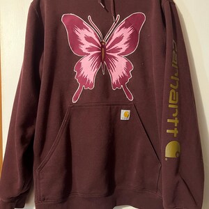 Puede incluir: Una sudadera con capucha Carhartt granate con un dise&ntilde;o de mariposa rosa. La sudadera tiene un bolsillo de canguro, una capucha con cord&oacute;n y el logo de Carhartt impreso en dorado en la manga.