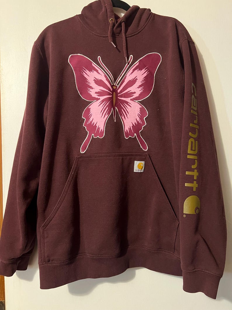 Puede incluir: Una sudadera con capucha Carhartt granate con un dise&ntilde;o de mariposa rosa. La sudadera tiene un bolsillo de canguro, una capucha con cord&oacute;n y el logo de Carhartt impreso en dorado en la manga.