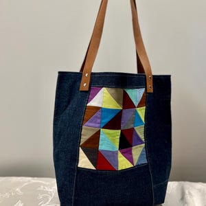 May include: A denim tote bag with brown leather handles. The front features a patchwork design of colorful geometric shapes in various colors, including blue, purple, yellow, and red. The bag is held up by a hand.