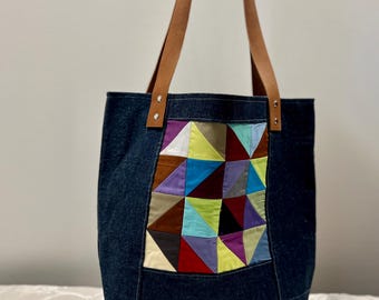 May include: A denim tote bag with brown leather handles. The front features a patchwork design of colorful geometric shapes in various colors, including blue, purple, yellow, and red. The bag is held up by a hand.