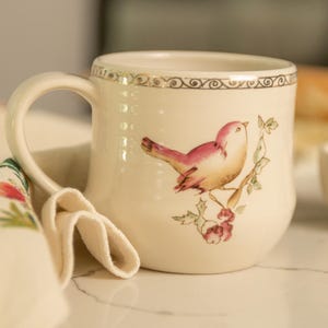 May include: A white ceramic mug with a gold rim and a painted design of a pink bird on a branch with green leaves and red berries.