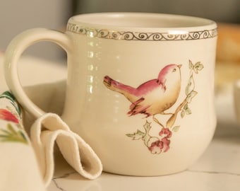 May include: A white ceramic mug with a gold rim and a painted design of a pink bird on a branch with green leaves and red berries.