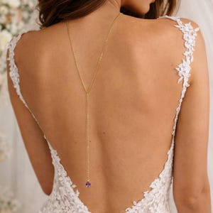 Op de afbeelding: Een goudkleurige Y-vormige lariat-ketting met een fijne ketting die langs de rug naar beneden valt en eindigt in een kleine, ronde, gefacetteerde paarse edelsteen hanger. De ketting wordt gedragen bij een mouwloze witte kanten jurk.
