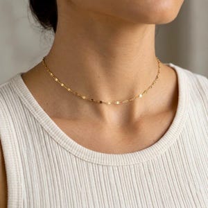 Könnte beinhalten: Eine zarte Goldkettenhalskette mit kleinen, flachen, reflektierenden ovalen Gliedern, die einen schimmernden Effekt um den Hals erzeugen. Die Kette wird über einem weißen gerippten Oberteil getragen und betont den metallischen Glanz der Goldglieder.