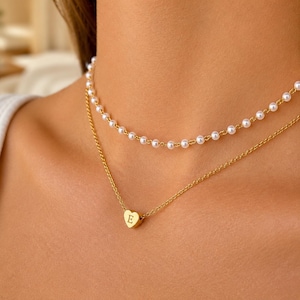 Peut inclure: Un ensemble de colliers superposés composé d'un rang de petites perles blanches rondes et d'une chaîne dorée avec un petit pendentif en forme de cœur doré gravé de la lettre majuscule E au centre de la chaîne.