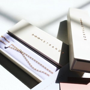 May include: A delicate rose gold chain necklace rests inside an open rectangular cream-colored box. The box lid features the text BODY STRANDS in metallic rose gold lettering. A second matching box stands vertically nearby with the same branding.