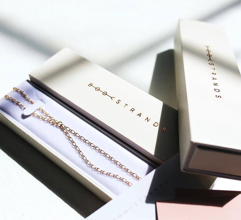 May include: A delicate rose gold chain necklace rests inside an open rectangular cream-colored box. The box lid features the text BODY STRANDS in metallic rose gold lettering. A second matching box stands vertically nearby with the same branding.