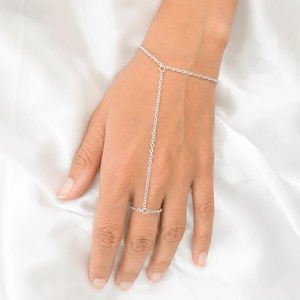 Puede incluir: Una pulsera de mano plateada que presenta una delicada cadena de eslabones metálicos que rodea la muñeca y se extiende por el dorso de la mano para conectarse a un anillo que rodea el dedo medio con un diseño de cadena simple.