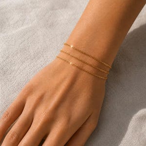 May include: Three delicate gold chain bracelets are worn on a wrist. The bracelets are thin and simple, with a subtle shine. The wrist is positioned over a neutral-colored fabric, creating a soft background. The bracelets are a minimalist accessory.