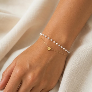 Op de afbeelding: Een dubbelstrengs armband bestaande uit een fijne goudkleurige ketting met een kleine hartvormige bedel gegraveerd met de letter E, en een tweede streng bestaande uit kleine, uniforme witte parels op een dunne ketting.