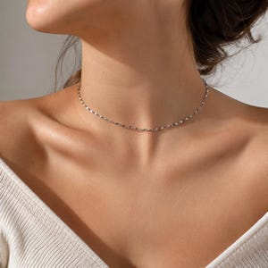 May include: A delicate, thin silver-toned chain necklace worn as a choker. The necklace features small, reflective, oval-shaped metallic links that catch the light, creating a subtle shimmering effect around the base of the neck.