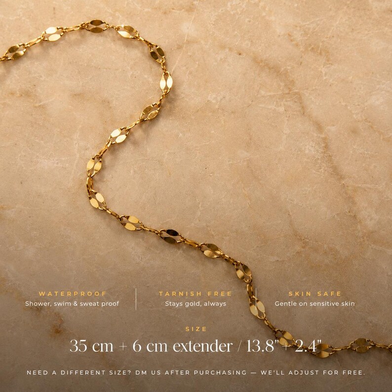 May include: A gold-toned chain necklace featuring elongated, flattened oval links. Text on the image specifies: WATERPROOF, TARNISH FREE, SKIN SAFE, and a size of 35 cm + 6 cm extender / 13.8" + 2.4". The text also includes a note about size adjustments.