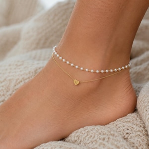 May include: A double-layered anklet featuring one strand of small white pearls and a second thin gold-toned chain with a small heart-shaped charm centered on the chain. The heart charm is engraved with the capital letter E in a serif font.