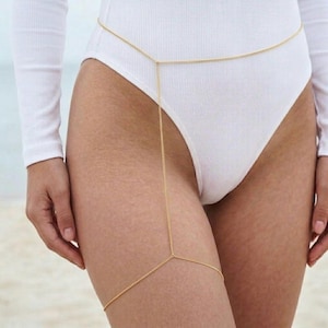 May include: A gold body chain. The chain is worn around the waist and extends down the leg. The chain is made of small, delicate links. The person is wearing a white bodysuit.