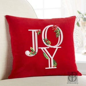 May include: A red decorative pillow with the word "JOY" embroidered in white. The letters are adorned with festive holly, berries, and pine needles, creating a holiday theme. The pillow is square and rests on a cream-colored surface.