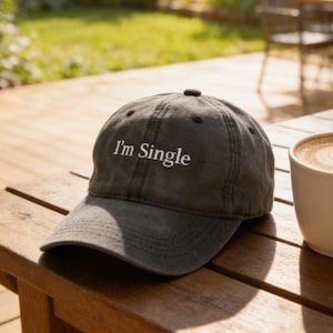 Könnte beinhalten: Eine graue Baseballkappe mit dem Text "I'm Single" in weißen Buchstaben liegt auf einem Holztisch neben einer weißen Kaffeetasse mit Latte Art. Die Szene spielt sich im Freien mit verschwommenem Hintergrund ab.