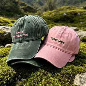 May include: A distressed olive green baseball cap with the embroidered pink text "Don't seduce me!" and a distressed light pink baseball cap with the embroidered dark green text "Süssmaus", both featuring a curved brim and stitched eyelets.