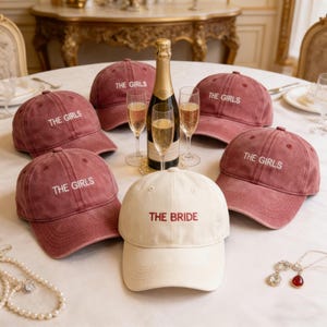 Peut inclure: Un ensemble de cinq casquettes de baseball disposées sur une table. Quatre casquettes sont rose poudré avec le texte THE GIRLS brodé en blanc. Une casquette centrale est blanc cassé avec le texte THE BRIDE brodé en rouge. Une bouteille de champagne et deux verres sont présents.