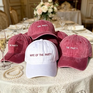 May include: A selection of baseball caps, predominantly in a faded red colour, with one white cap. The red caps are embroidered with "THE PARTY", while the white cap reads "WIFE OF THE PARTY". A pearl necklace and champagne glass are also visible.