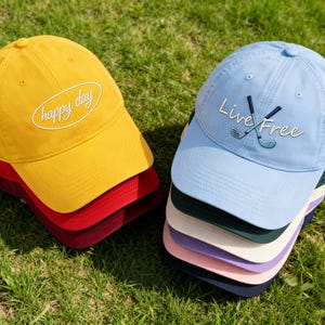 Puede incluir: Dos gorras de béisbol apiladas sobre hierba. Una es amarilla con "happy day" bordado en blanco. La otra es azul claro con "Live Free" y bordado de palos de golf. Se ven gorras adicionales en rojo, burdeos, verde, blanco, rosa y marino.
