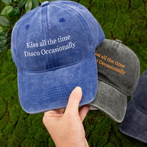 Könnte beinhalten: Blaue und graue Baseballkappen mit dem Text "Kiss all the time Disco Occasionally". Die blaue Kappe wird von einer Hand gehalten. Der Hintergrund ist grün und moosig. Lässiges Modeaccessoire.